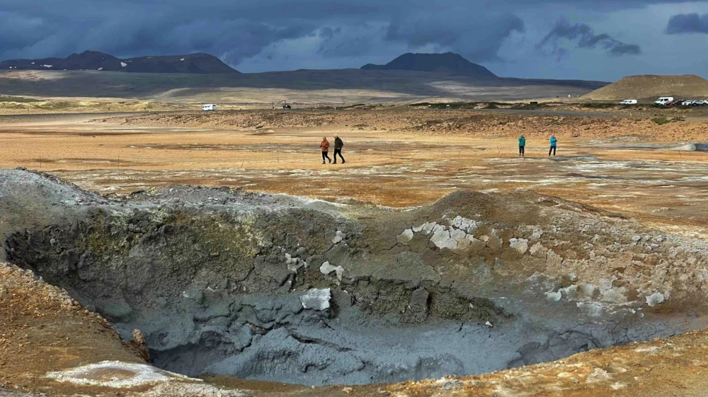 tourists exploring a geothermal area with boiling mud pools and steam vents, set against a backdrop of a dramatic sky and distant mountains