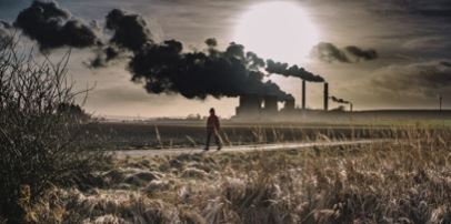 A person walking in between two fields with a factory in the background