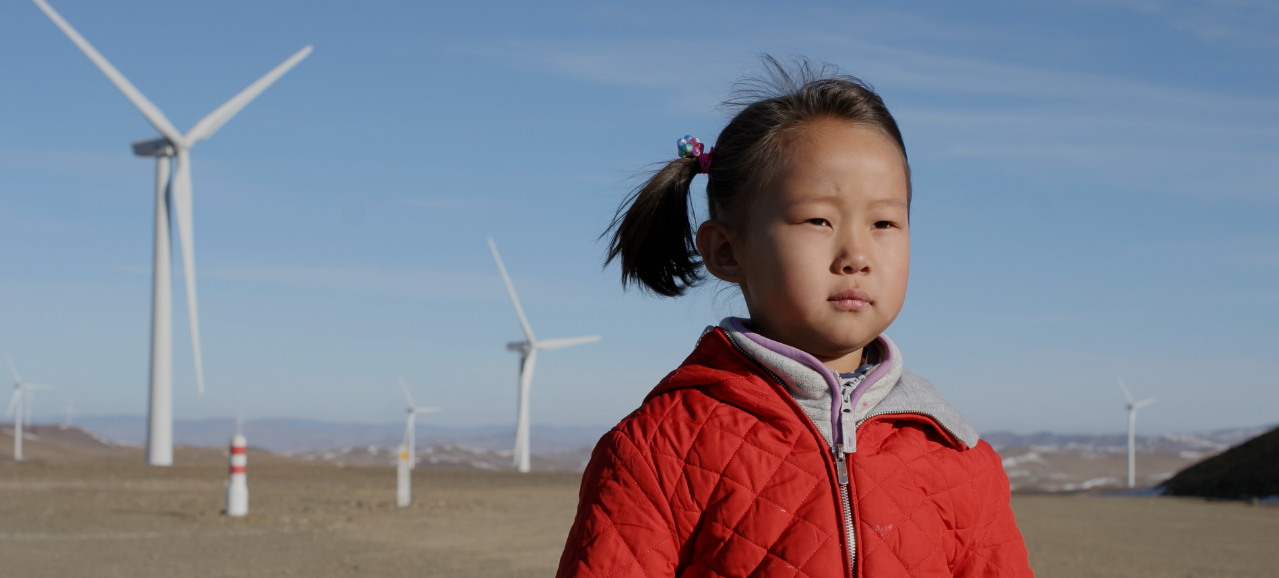 Young girl in red jacket stands among turbines at Salkhit Wind Farm in Mongolia