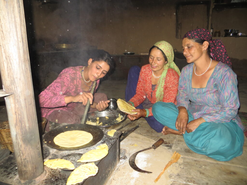 Three women sitting around a stove, one blowing through a pipe to stoke the fire.