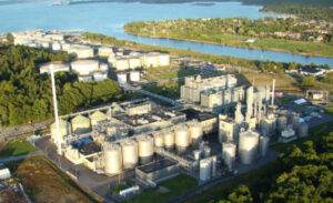 Aerial view of an industrial site with lots of tanks and processing equipment. It is surrounded by trees and a river meets the sea in the background.
