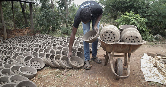 Person arranging rows of clay cooking stoves beside a wheelbarrow outdoors.