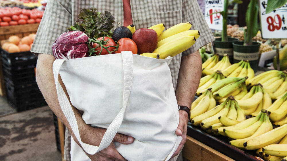 Bananas, cabbage and other fruits and veg in a cloth bag held by a man.