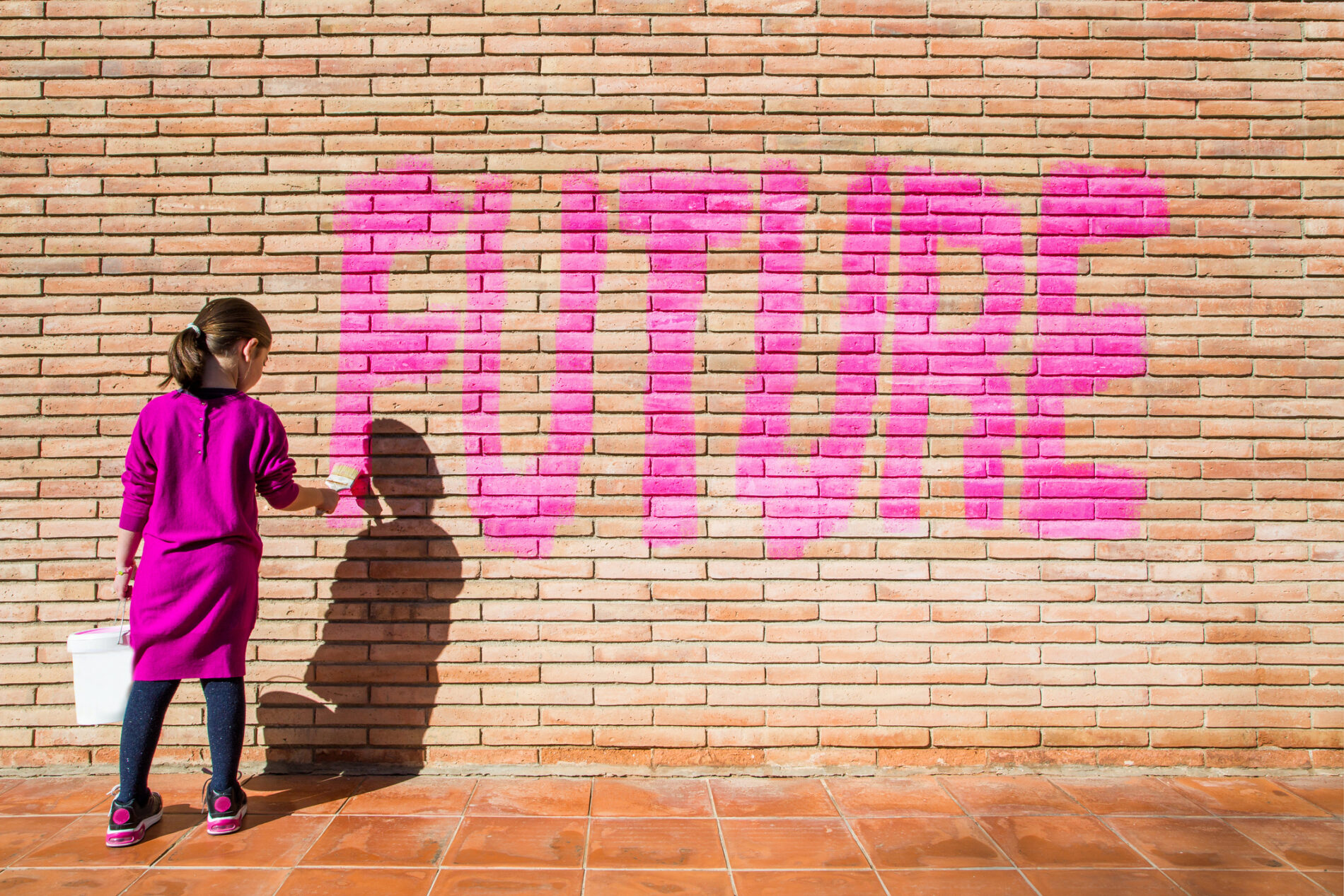 Little girl painting Future in pink on brick wall.