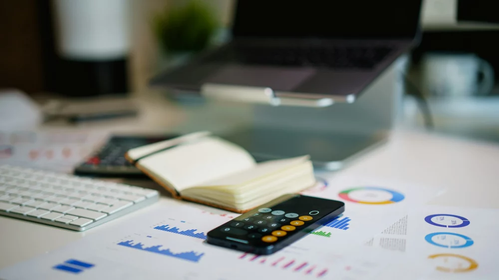 desk with laptop, papers, calculator app on phone
