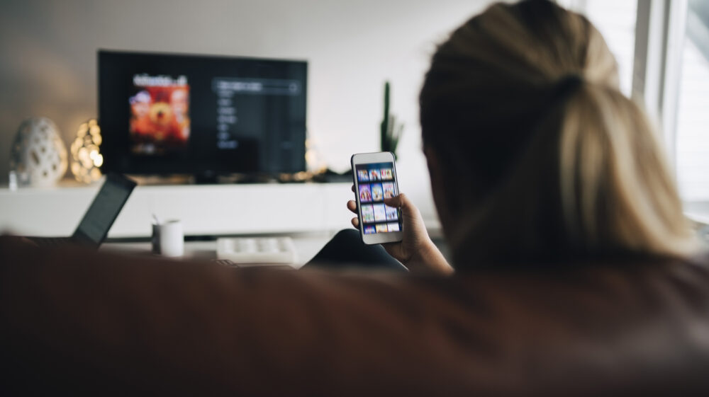 Rear view of teenage girl using smart phone app while watching TV in living room at home