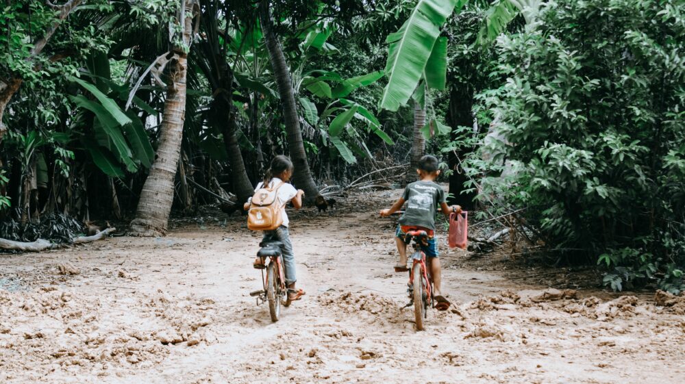 Girl wearing a backpack with a fake APPLE logo and boy with pink plastic bag on right handlebar ride bikes to school on unpaved path in forest to school, Paoy Paet, Krong Poi Pet, Cambodia
