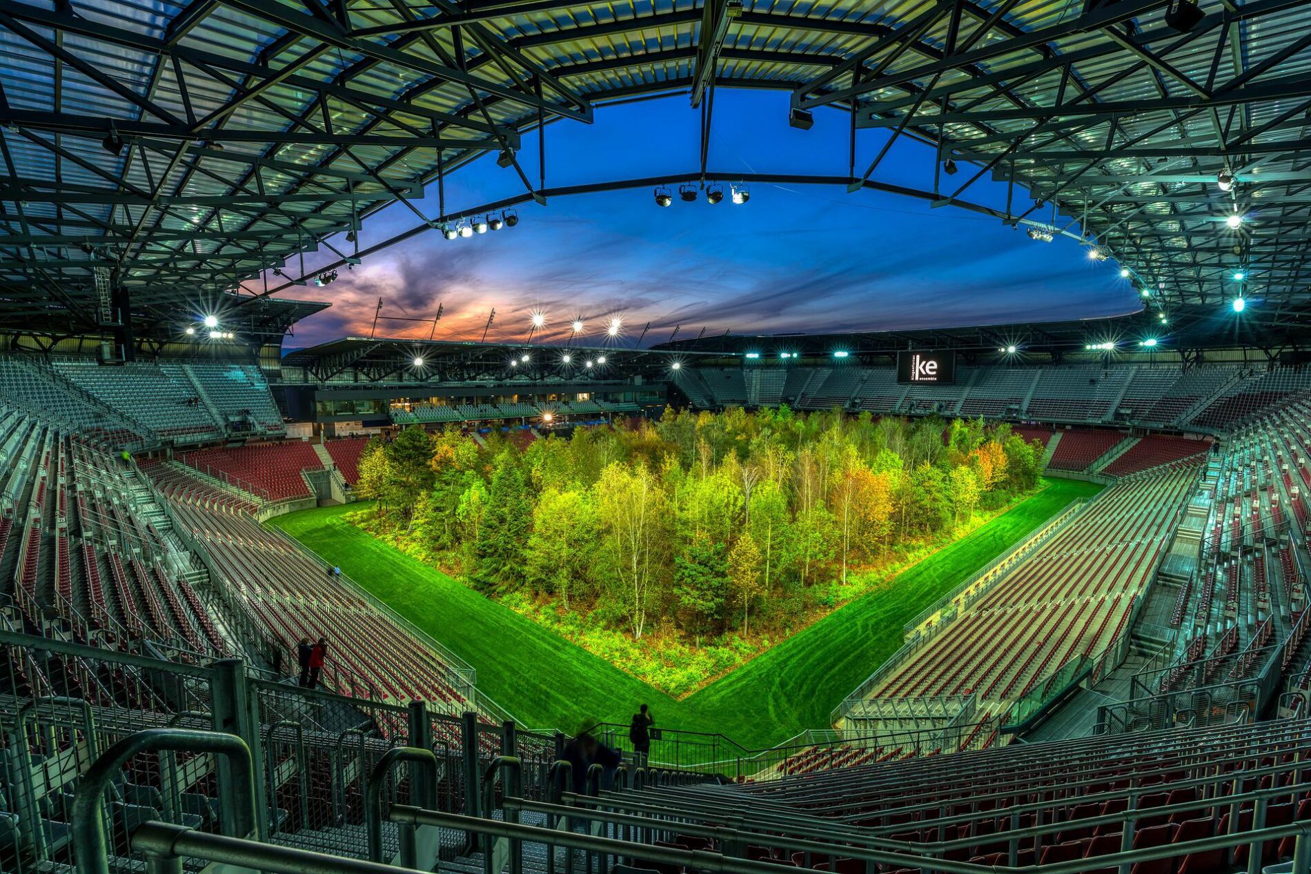 Composite of green forest in stadium at dusk under light purple and blue sky and stadium lights, Klagenfurt, Austria