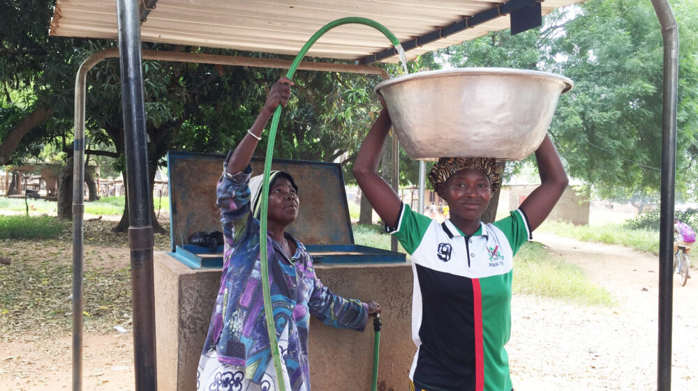 An African woman fills a large metal tub in front of her above another African woman's head using a water hose