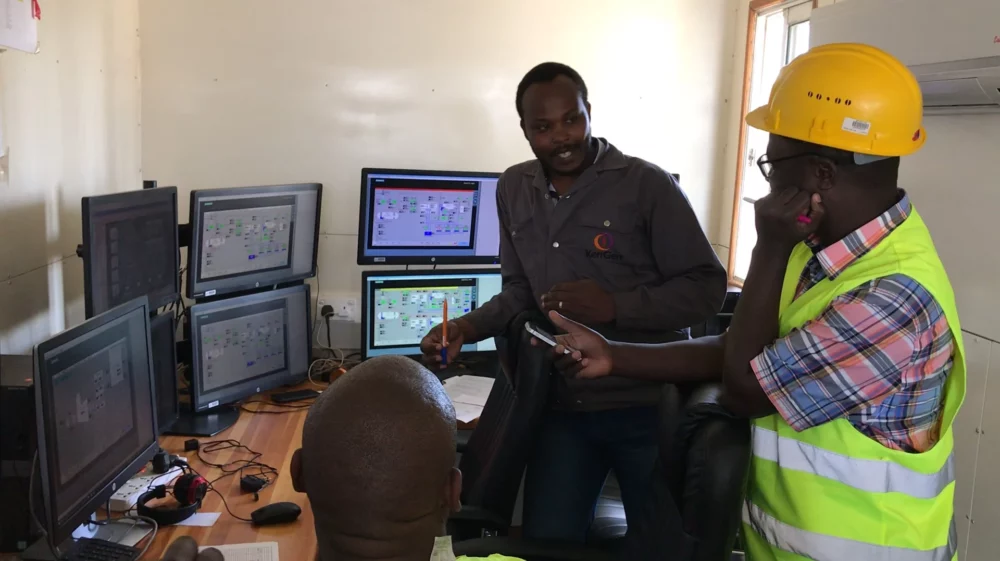 Engineers collaborating in a control room, with multiple computer monitors displaying technical data, one wearing a yellow safety helmet and vest.
