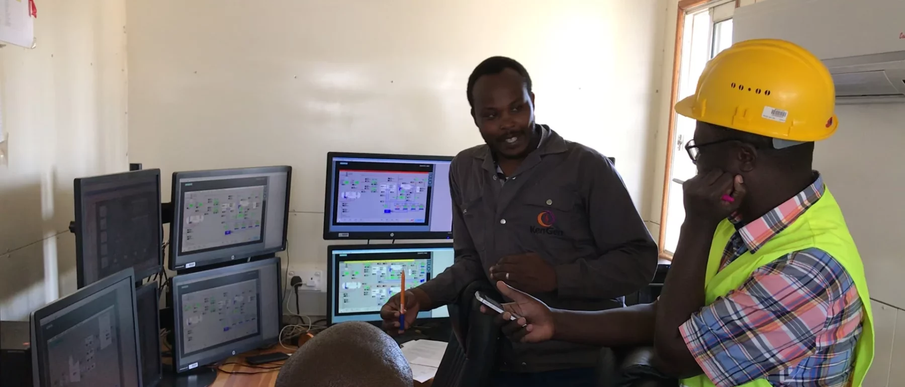 Engineers collaborating in a control room, with multiple computer monitors displaying technical data, one wearing a yellow safety helmet and vest.