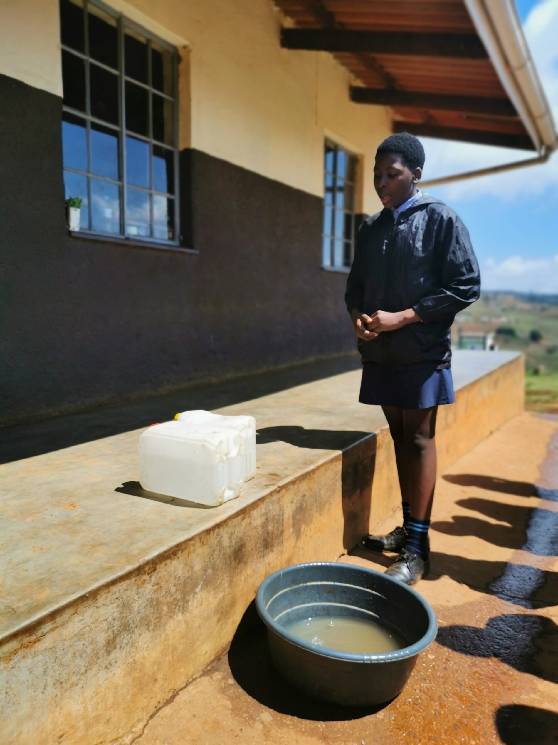 A student on schools grounds with a bucket grey water and a jerrycan with fresh water.