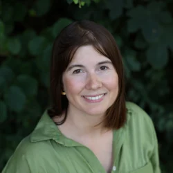 Head shot shows Charlotte wearing a green collared top smiling up at the camera with green foliage in the background