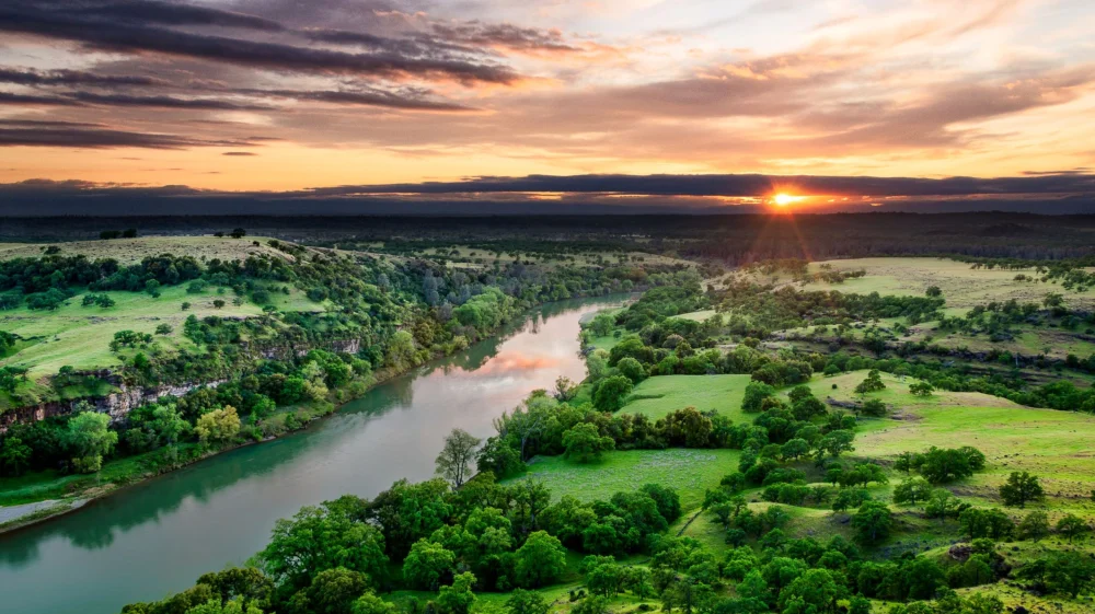 Sacramento River in California cuts through green tree-covered grassland at sunset.