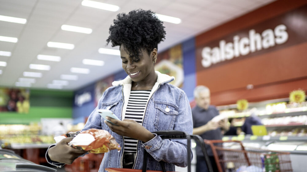 Black woman holding grocery product in supermarket in Brazil while holding phone in other hand