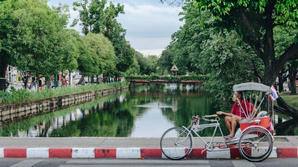 Male rickshaw driver waits for customers next to canal enveloped by green trees during afternoon, Chiang Mai, Thailand