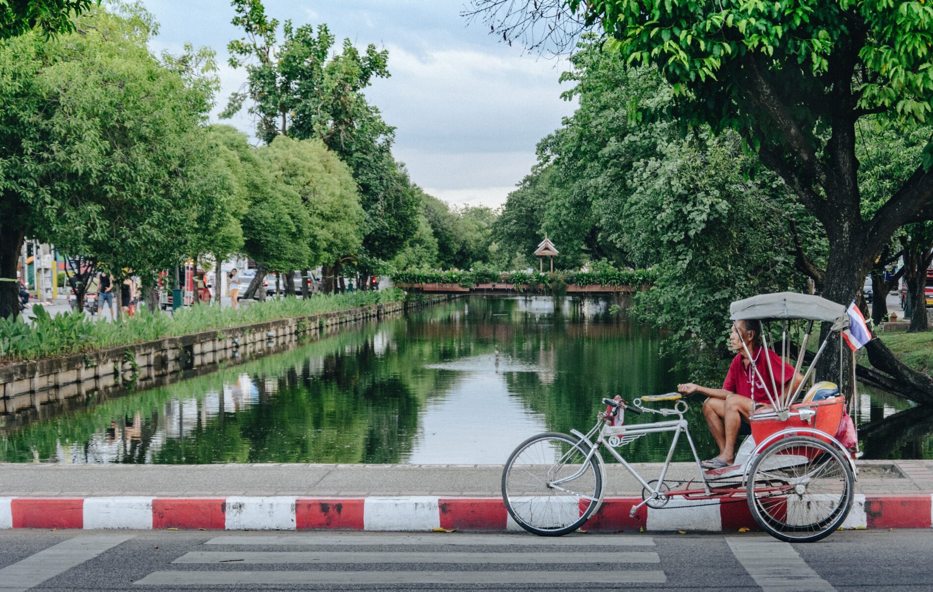 Male rickshaw driver waits for customers next to canal enveloped by green trees during afternoon, Chiang Mai, Thailand