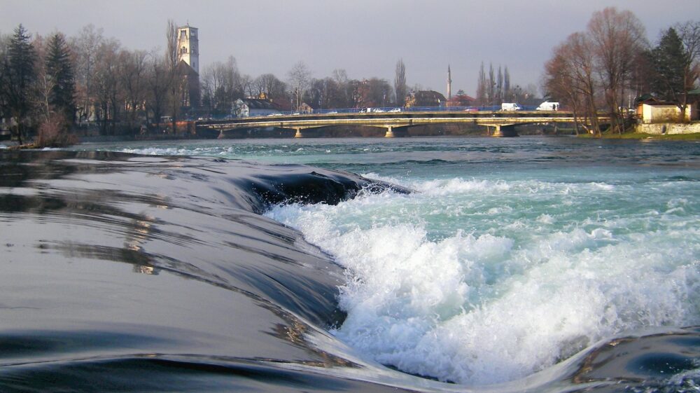 Cascade on Una River with bridge in background snd church and minaret further in distance, Bihać, Bosnia and Herzegovina