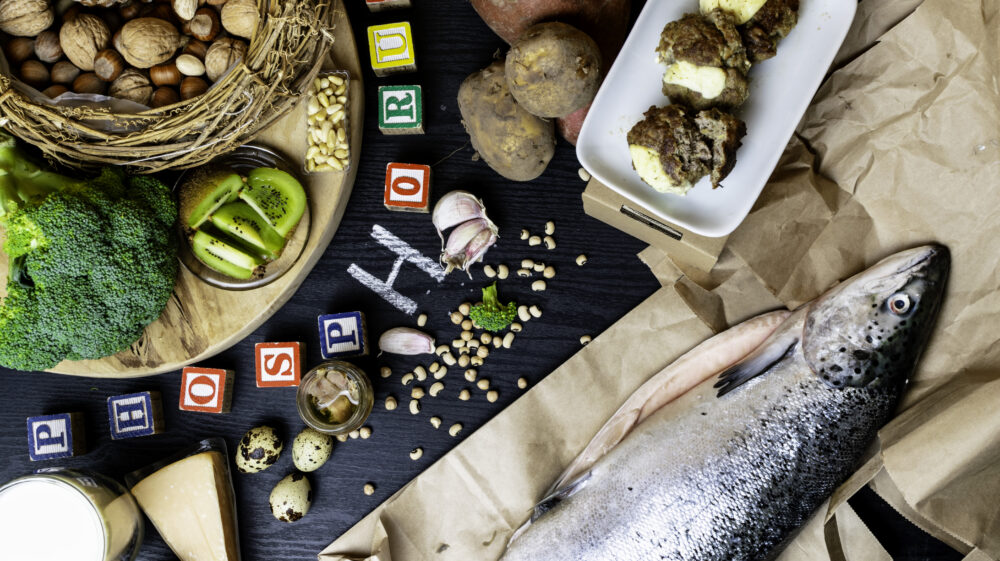 Products containing phosphorus salmon fish, pine nuts, peanuts, anchovy, cheese, milk, eggs, broccoli, kiwi, potatoes, meat, beans garlic on a round cutting board and black wooden background