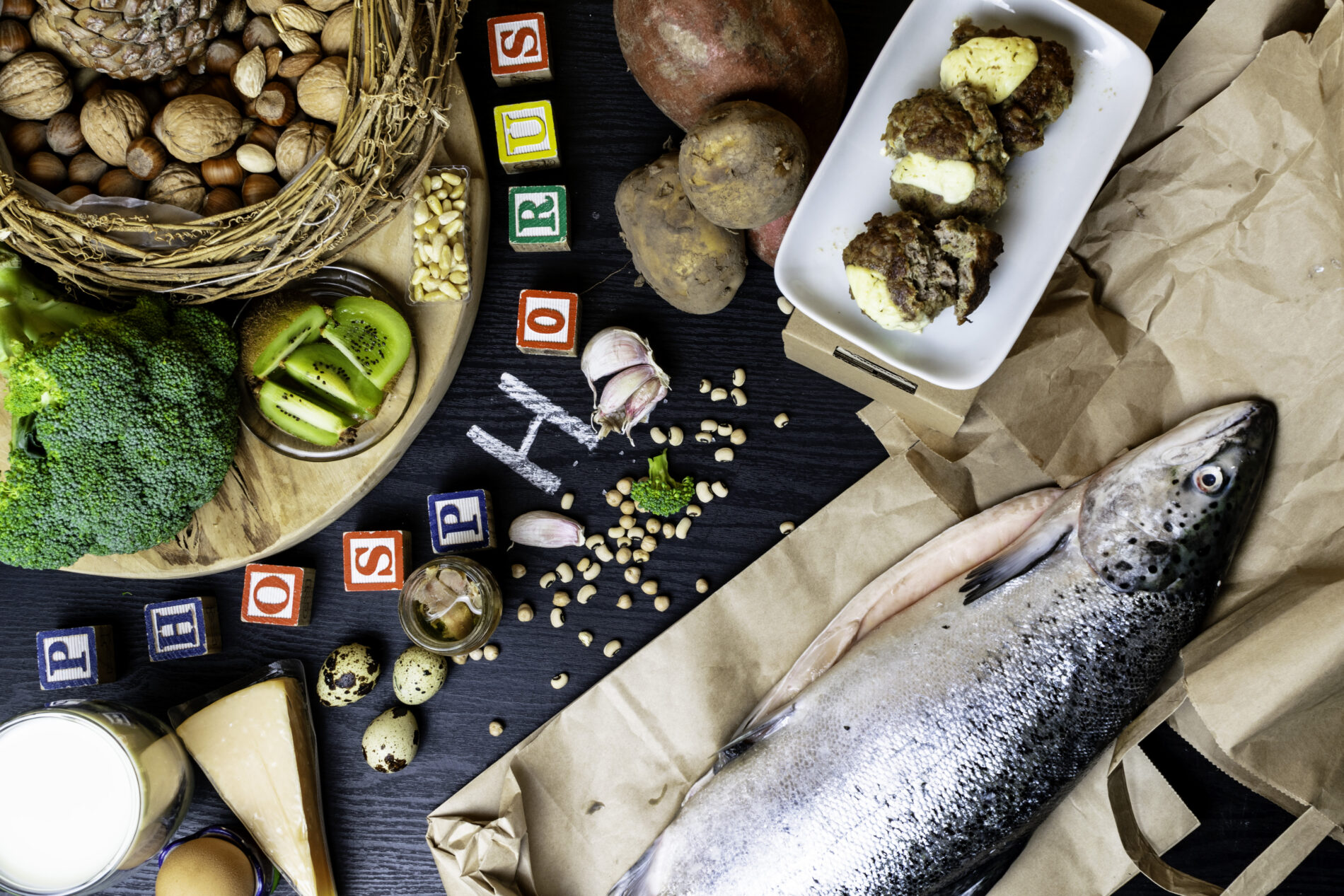 Products containing phosphorus salmon fish, pine nuts, peanuts, anchovy, cheese, milk, eggs, broccoli, kiwi, potatoes, meat, beans garlic on a round cutting board and black wooden background