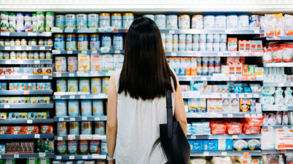 Rear view of young Asian mother groceries shopping for baby products in a supermarket. She is standing in front of the baby product aisle and has no idea which product to choose from