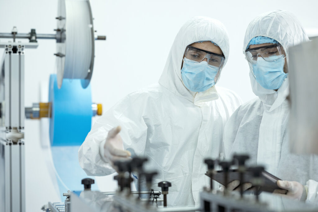 Two workers in full personal protective equipment at a factory looking over equipment, one holding a digital tablet