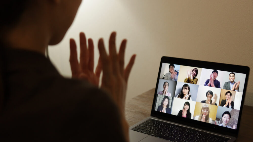 Five Japanese men and seven Japanese women on a screen clapping during a web conference along with woman watching on laptop, Japan