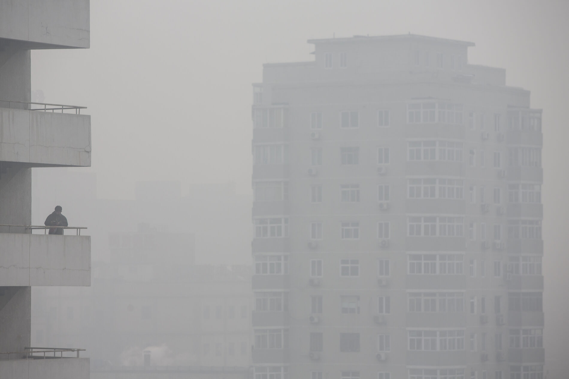 A man stands on a balcony as buildings stand shrouded in haze in Beijing, China