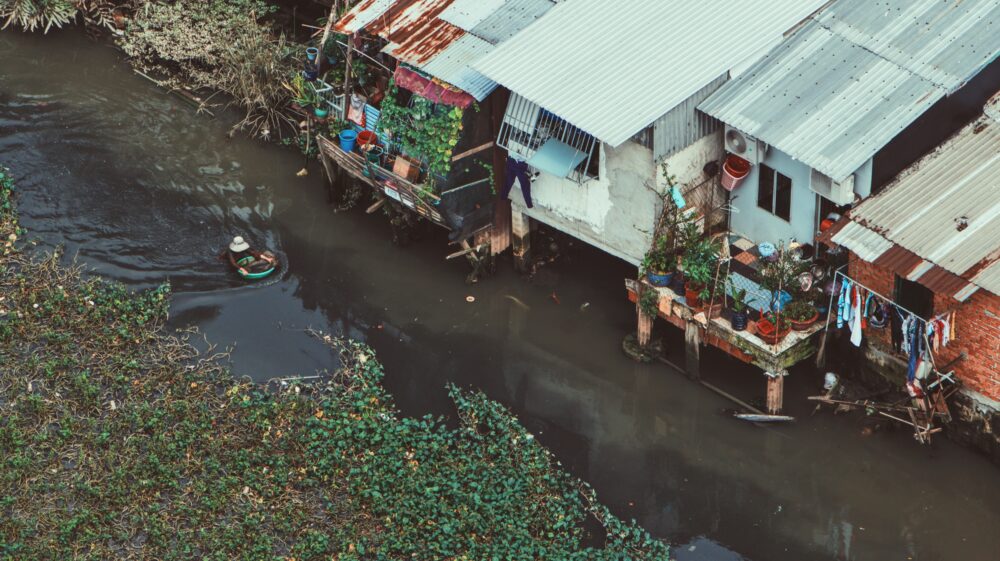 Woman swimming in canal with goods next to homes on stilts, Ho Chi Minh City, Viet Nam