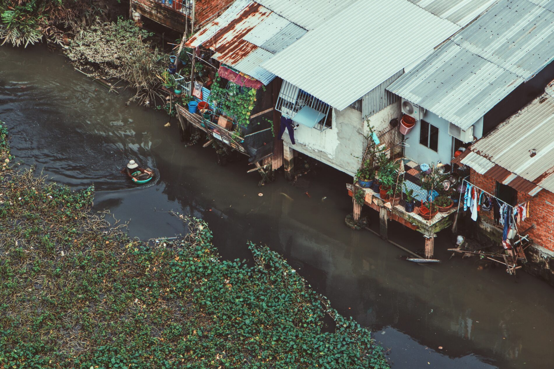Woman swimming in canal with goods next to homes on stilts, Ho Chi Minh City, Viet Nam