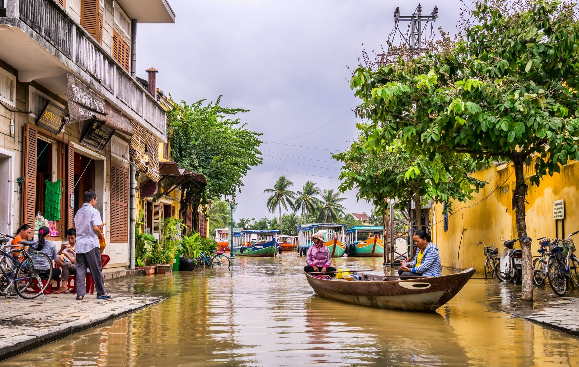 Two women in rowboat on canal between shophouses flooded up to sidewalks during rainy season on overcast day, Hoi An, Viet Nam