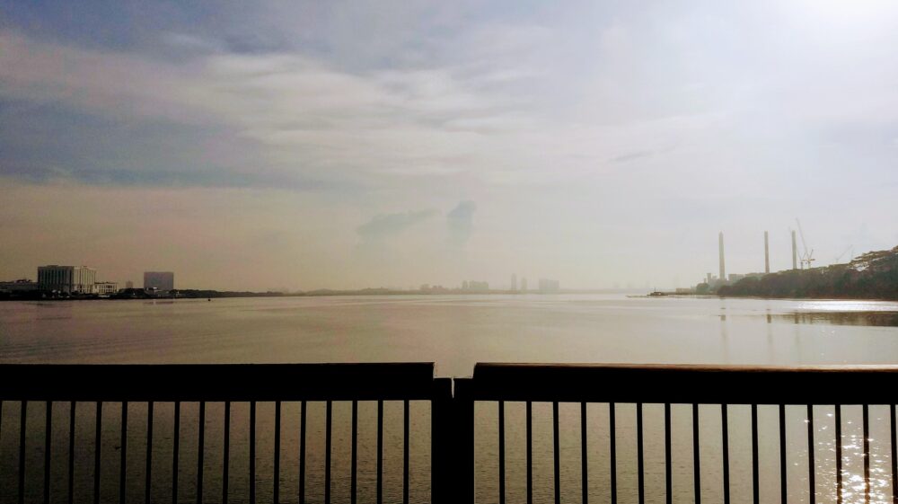 Black wooden handrail next to body of water with buildings in the distance on hazy day