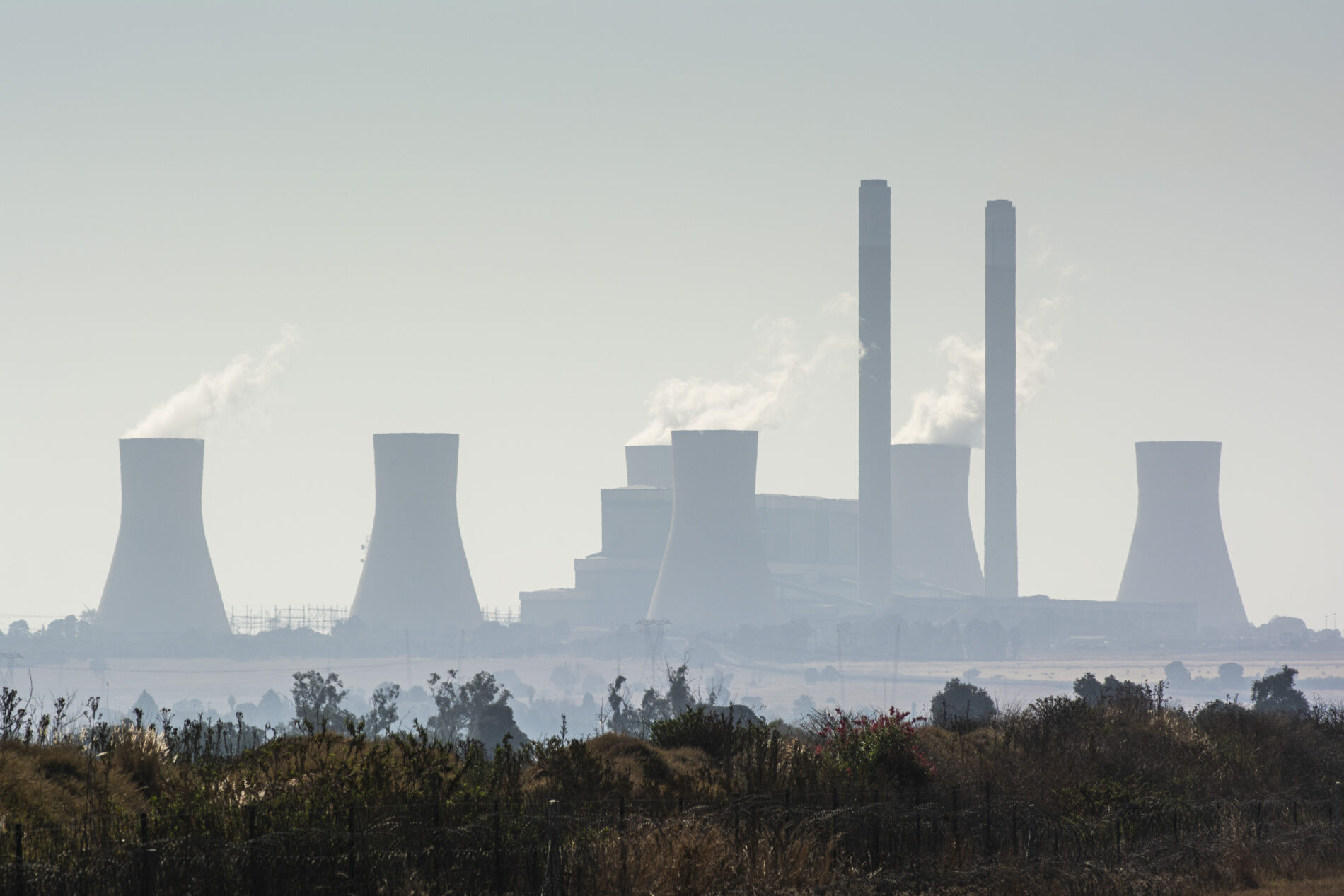 Vapour rises from cooling towers in distance on hazy day with grass in front, Mpumalanga, South Africa