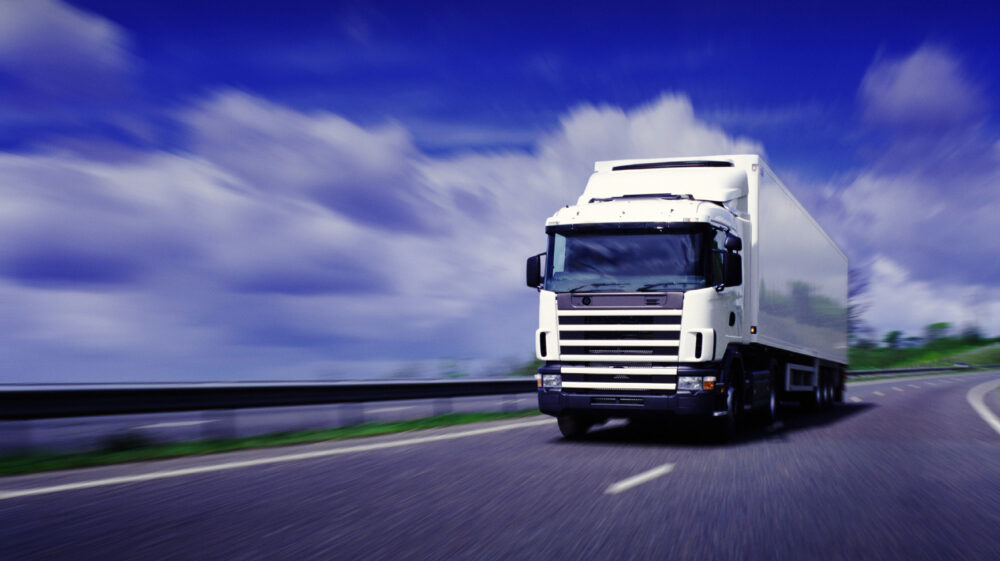 Lorry driving on motorway. Digital enhancement with clouds in blue skies, guard rail, road and green grass faded out to focus on lorry