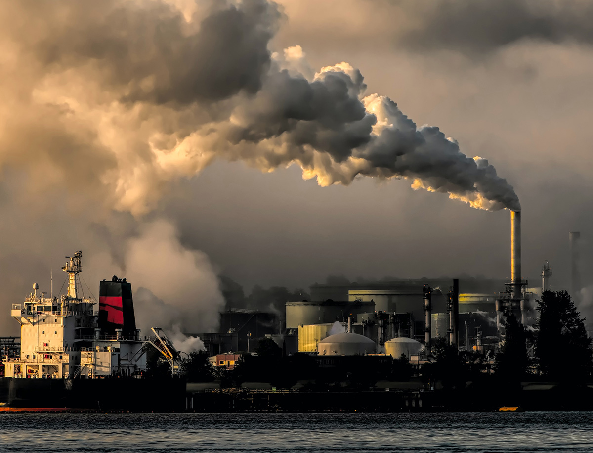 White ship with black and red mast docked at factory under billowing clouds from smokestack
