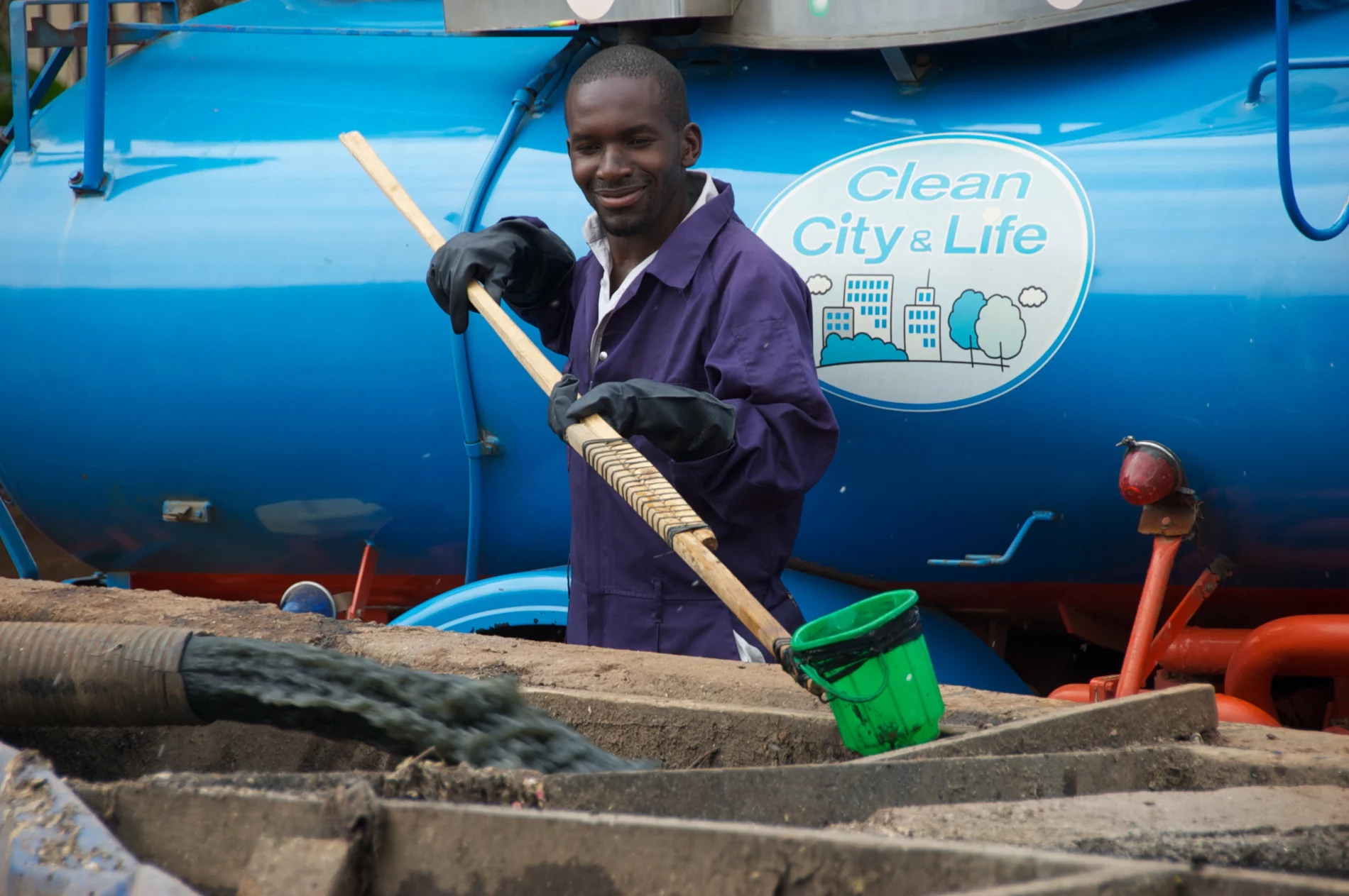 Daniel Ddiba sampling wastewater in front of a blue sanitation truck with a ‘Clean City & Life’ logo.
