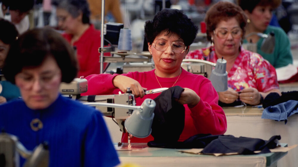 Women working in garment industry sit in rows behind sewing machines and lamps using scissors to cut fabric