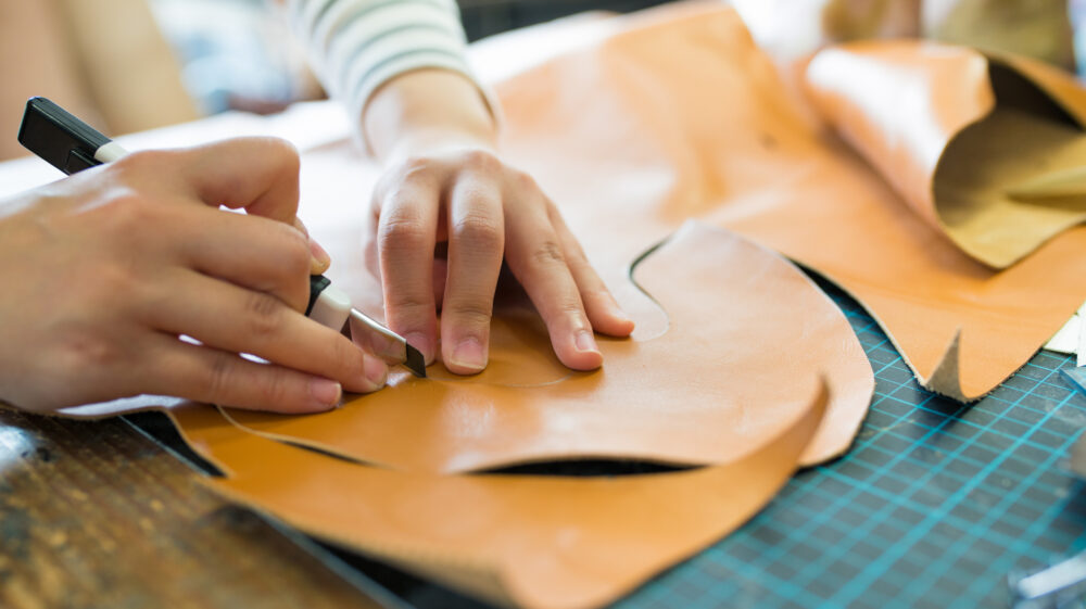 The photo shows a tabletop mostly covered with light brown leather being cut by a pair of hands holding a special knife.