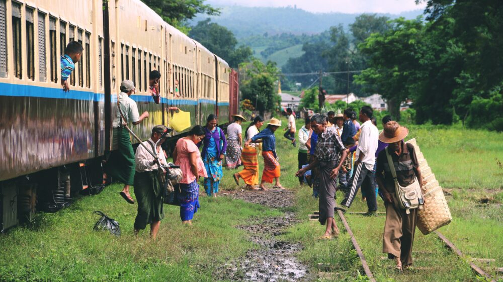 Passengers disembarking train onto green grass and mud with green hills and green trees in distances, Hsipaw, Myanmar, March 2018