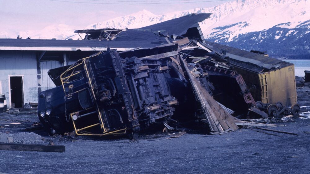 Truck on side outside damaged house by tornado, mountains in the background, wrecked black car on grey sand, Alaska 1964 Good Friday earthquake and tsunami damage