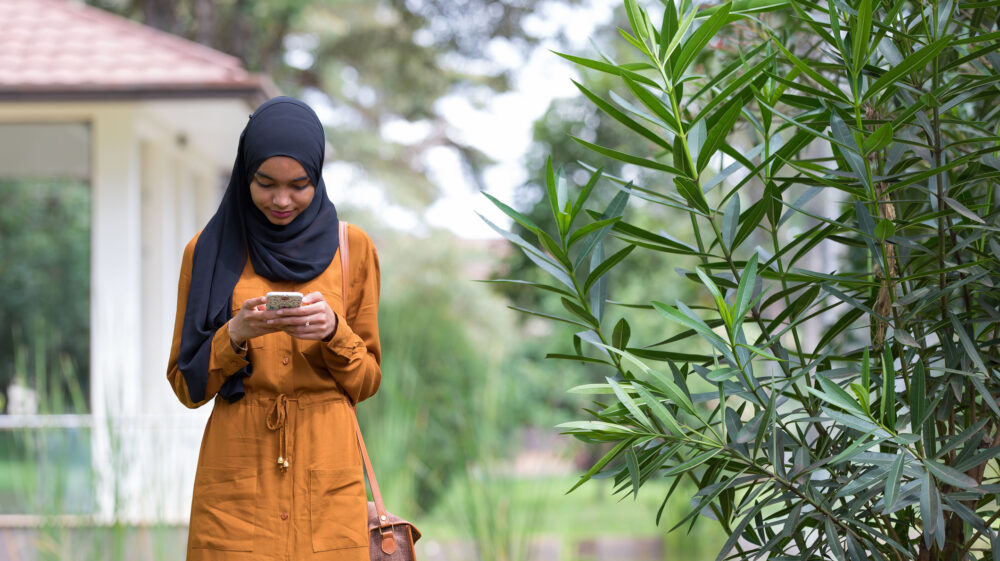 Woman in black hijab and orange dress with long-strapped shoulder bag texting on phone with red-roofed white pavilion behind her standing next to green plant, Kenya