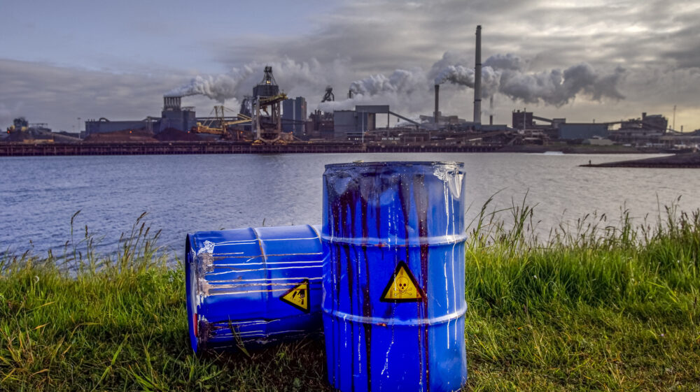 Empty blue chemical waste drums lie on abandoned bank with view of smoking exhaust Pipes from heavy industry factory, the Netherlands