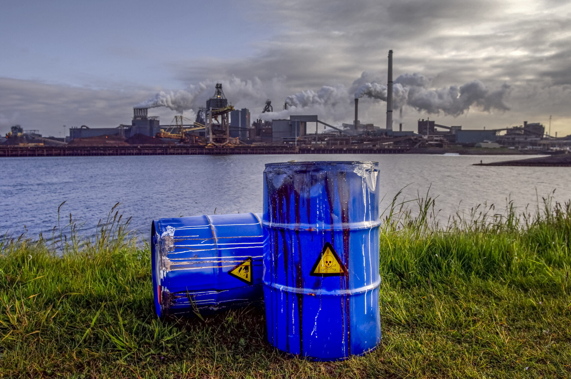 Empty blue chemical waste drums lie on abandoned bank with view of smoking exhaust Pipes from heavy industry factory, the Netherlands