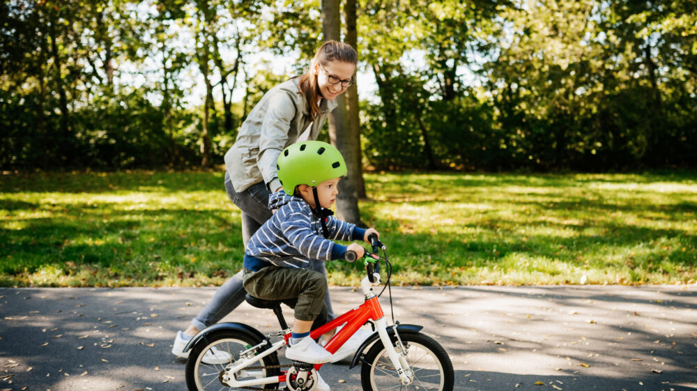 Mom guiding young son wearing lime green helmet and learning to ride red bike in park, Berlin, Germany