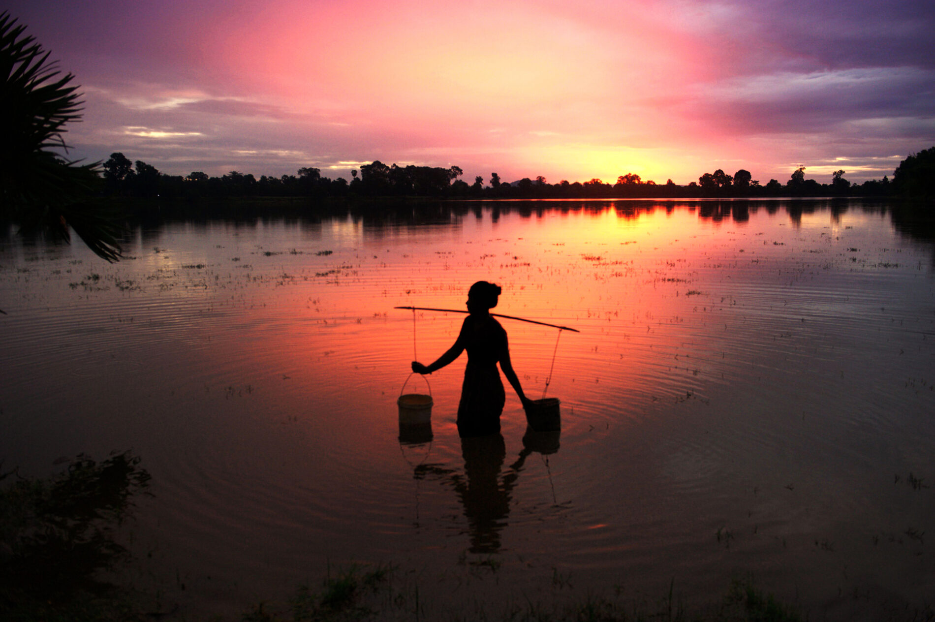 Girl in Sra Sang collecting water at dusk under yellow, pink and purple skiy in ancient bathing pool in countryside, Siem Reap, Cambodia
