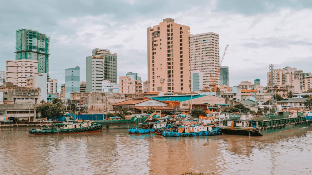 Blue and white boats on muddy water near tan and green buildings under cloudy sky, Fort Santiago, Intramuros, Manila, Philippines, February 2021