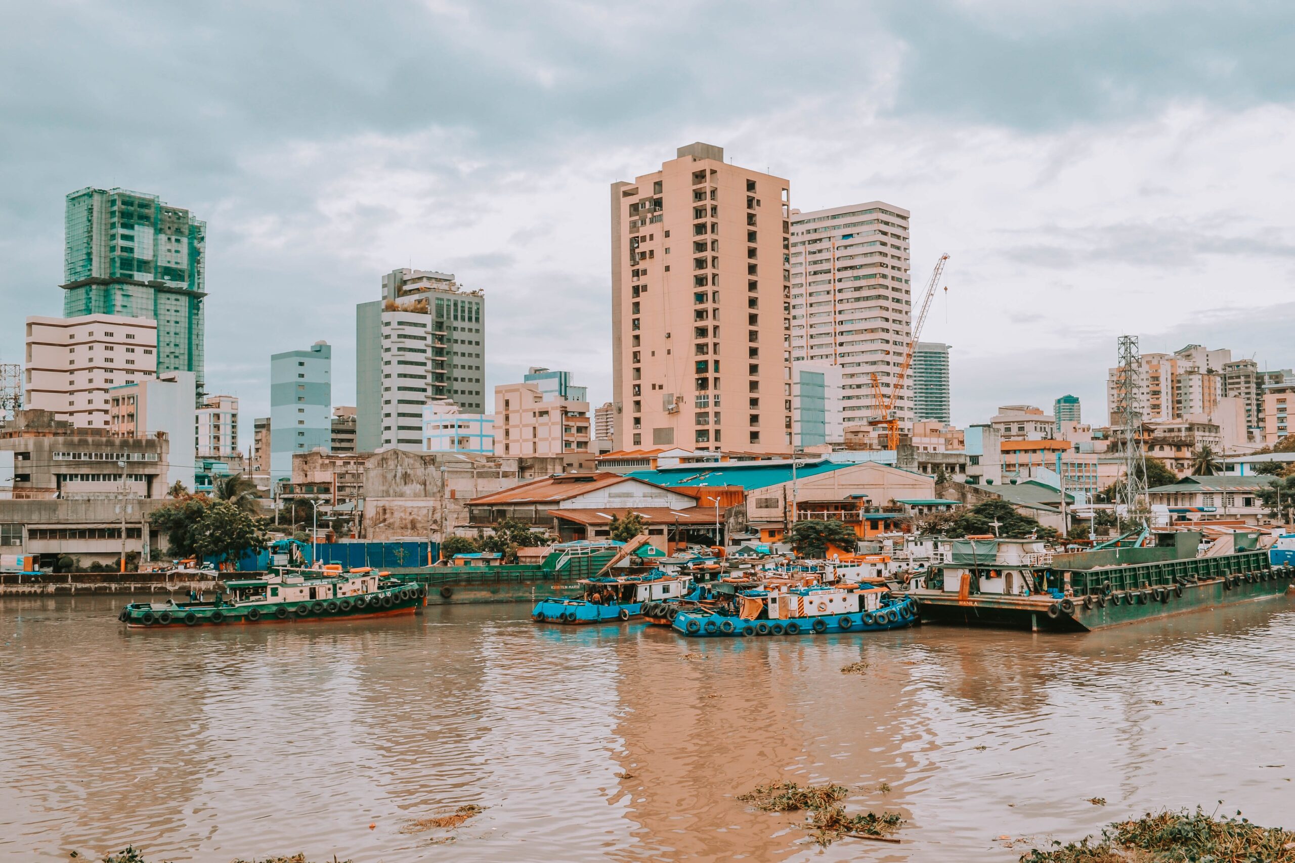 Blue and white boats on muddy water near tan and green buildings under cloudy sky, Fort Santiago, Intramuros, Manila, Philippines, February 2021