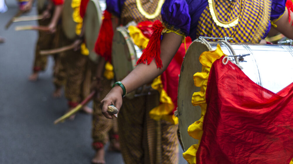 Indian women in purple, yellow and red costumes drumming in parade with red and yellow-trimmed coverings on drums, Mangalore, Karnataka, India
