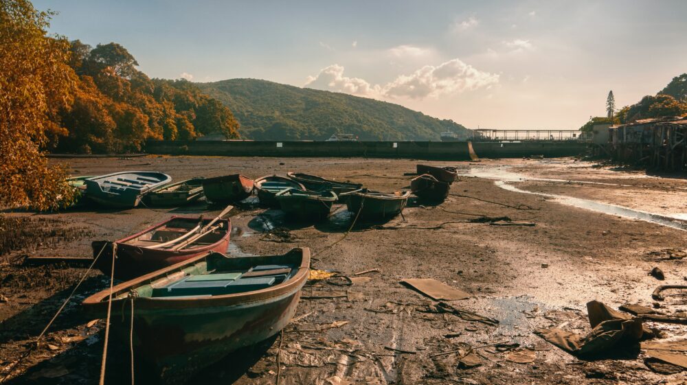 Green, red and blue rowboats on dried-out seabed with trees and mountain in background, Ma Wan, Hong Kong, August 2016