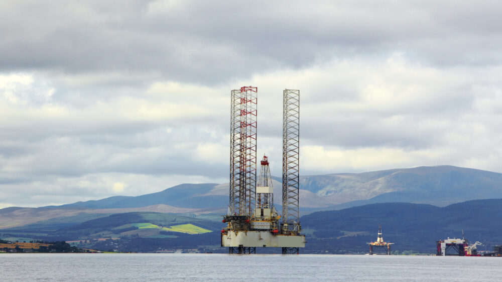 Jackup rig used for offshore oil and gas drilling stands under overcast sky in Scotland with rolling green hills in background, Cromarty Firth, Ross and Cromarty, UK